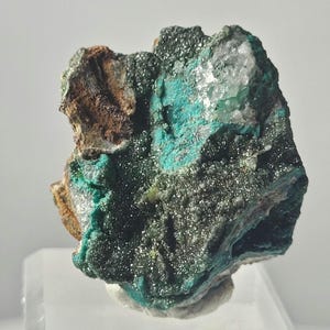 May include: A close-up of a mineral specimen featuring a mix of colors and textures. The mineral displays a vibrant turquoise hue, contrasted by areas of dark green, brown, and sparkling silver. The rough, irregular shape suggests a natural, unpolished state.