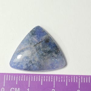 K&ouml;nnte beinhalten: Ein dreieckiger, polierter Edelstein mit einem blau-grauen, melierten Muster. Der Stein hat eine glatte, gl&auml;nzende Oberfl&auml;che und ist etwa 2,5 cm breit. Der Edelstein liegt vor einem wei&szlig;en Hintergrund, mit einem lila Lineal im Vordergrund.