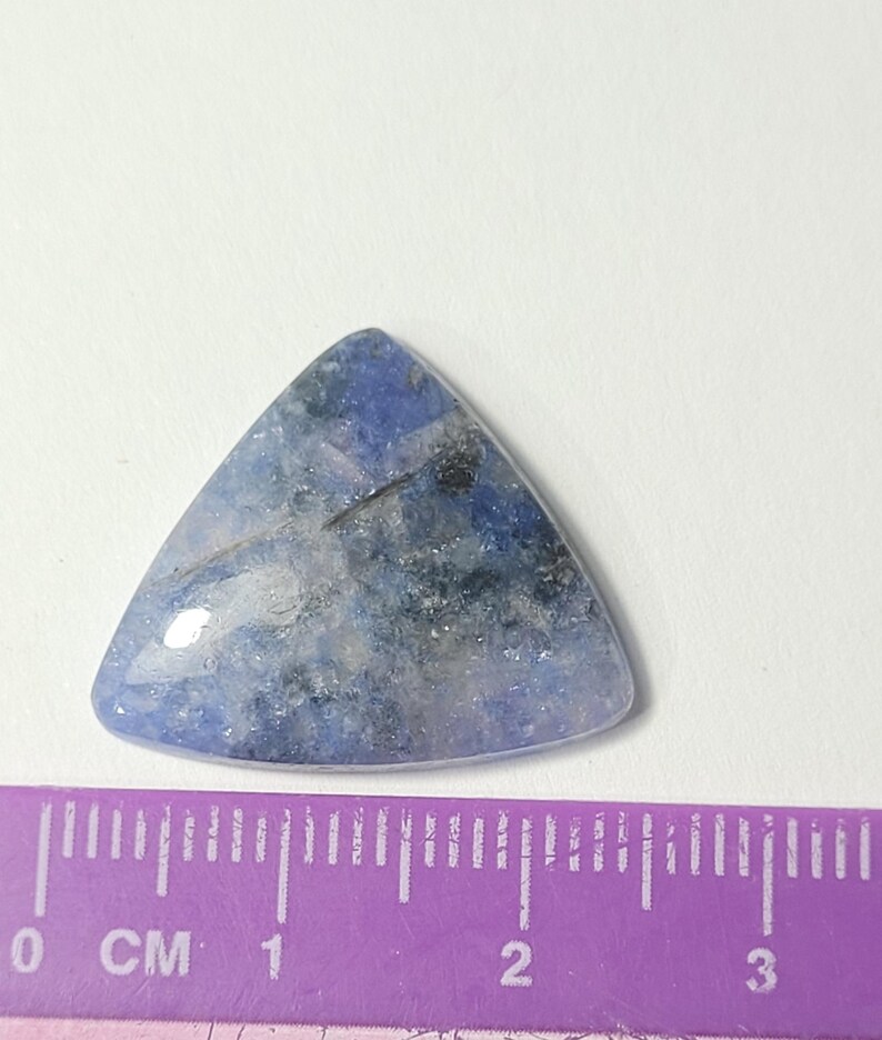 K&ouml;nnte beinhalten: Ein dreieckiger, polierter Edelstein mit einem blau-grauen, melierten Muster. Der Stein hat eine glatte, gl&auml;nzende Oberfl&auml;che und ist etwa 2,5 cm breit. Der Edelstein liegt vor einem wei&szlig;en Hintergrund, mit einem lila Lineal im Vordergrund.