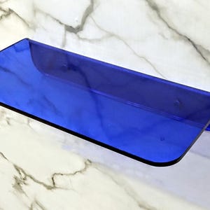 May include: A rectangular wall-mounted shelf made of translucent cobalt blue acrylic. The shelf features a flat horizontal surface with a curved upward-facing edge along the back, providing a sleek and simple design for storage or display purposes.