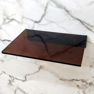 May include: A rectangular shelf made of dark brown, semi-transparent glass. The shelf features a smooth, flat surface with clean, sharp edges and is mounted horizontally, showcasing a deep, reflective, and uniform amber-toned glass construction.