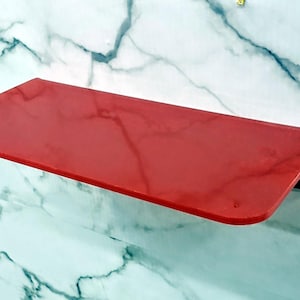 Könnte beinhalten: Ein rechteckiges Wandregal mit einer glatten, einfarbig roten Oberfläche und abgerundeten vorderen Ecken. Das Regal ist an einer weißen Fläche mit grauer Marmorierung befestigt, was einen starken Kontrast für dieses Ablageelement erzeugt.