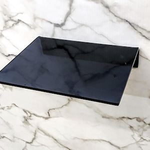 May include: A rectangular, solid black shelf with a smooth, reflective surface. The shelf features a clean, minimalist design with a single right-angle bend at the back edge, allowing it to be mounted flush against a flat vertical surface.
