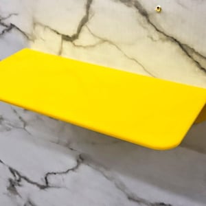 Può includere: Una mensola da parete rettangolare di colore giallo brillante con angoli arrotondati e una finitura liscia e solida. La mensola è montata orizzontalmente su una superficie bianca con venature di marmo grigio e una piccola vite metallica.