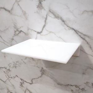 May include: A rectangular white marble shelf with a smooth, polished surface and straight edges. The shelf is mounted horizontally against a matching white marble wall featuring prominent grey veining patterns that run throughout the stone surface.