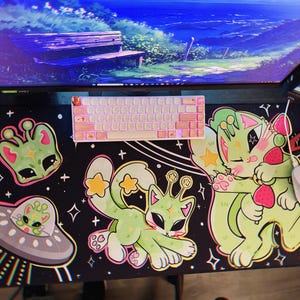 Puede incluir: Una alfombrilla de ratón negra con un diseño de temática espacial que presenta criaturas verdes parecidas a alienígenas, una nave espacial y estrellas. El diseño también incluye un gato rosa y verde con una fresa.