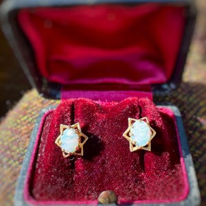 May include: A pair of gold-toned star-shaped earrings with oval, opalescent stones, displayed in a vintage black jewelry box with a red velvet interior. The earrings are the focal point, showcasing a classic design.