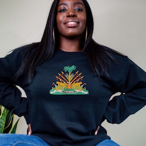May include: Black sweatshirt featuring a colorful embroidered emblem. The emblem includes a palm tree, cannons, and the text "L'UNION FAIT LA FORCE" in white. The sweatshirt is a solid black color.