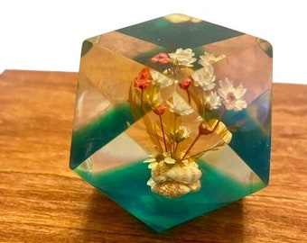 May include: A clear glass cube paperweight with a seashell and dried flowers inside. The flowers are white, pink, and red. The seashell is white and beige. The cube is filled with a teal blue liquid.