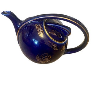 Puede incluir: Una tetera azul cobalto con un diseño floral dorado. La tetera tiene un cuerpo redondo y un asa curva.