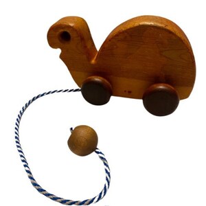 Puede incluir: Un juguete de madera con forma de tortuga con ruedas marrones y una cuerda azul y blanca. La tortuga tiene una bola de madera marrón en el extremo de la cuerda.