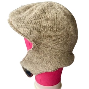 Puede incluir: Gorra de estilo newsboy tejida de color beige con orejeras. El gorro está hecho de un material texturizado similar a la lana. Las orejeras están diseñadas para brindar calidez y protección adicional contra el frío. La gorra se exhibe en una cabeza de maniquí rosa.