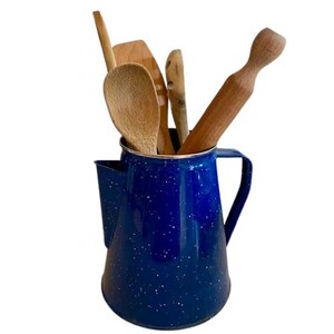Könnte beinhalten: Ein blauer Emaille-Krug mit Sprenkeln enthält Küchenutensilien aus Holz, darunter ein Löffel, ein Spatel und ein Nudelholz. Der Krug hat einen Griff und einen Ausguss. Die Utensilien sind aus hellem Holz.