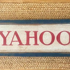 May include: Rectangular wooden sign with the word "YAHOO" in red letters. The sign has a distressed, weathered look with a blue frame and is set against a woven, tan background.