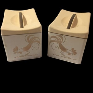 May include: Two vintage cream-colored metal canisters with rooster designs. Each canister has a brown plastic handle on the lid.