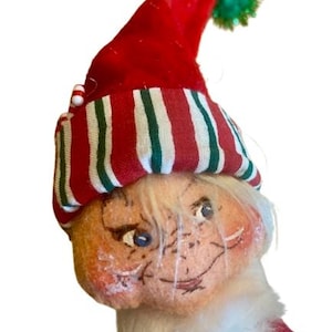 May include: A vintage Christmas elf doll with a red and white striped hat, a green pom-pom, and a white beard. The elf has a painted face with blue eyes and a mischievous smile.