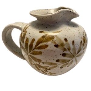 Può includere: Un'anfora in ceramica bianca con un motivo a foglie marroni. L'anfora ha una glassa macchiettata e un corpo arrotondato.