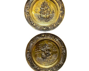 Op de afbeelding: Twee gouden metalen wandplaten met een sierlijk ontwerp. Elke plaat heeft een reliëfbeeld van een zeilschip met een gedetailleerd ontwerp.