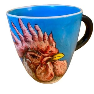 May include: A blue ceramic mug with a hand-painted rooster design. The rooster's head is facing the viewer, and its comb and wattle are a bright red. The mug has a black handle.