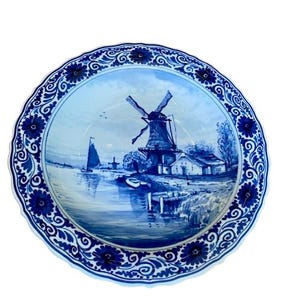 Könnte beinhalten: Eine runde, blau-weiße Zierteller mit einer holländischen Windmühlen-Szene. Der Teller hat einen gewellten Rand mit floralen und Rankenmustern. Die Windmühle, Boote und Wasser sind in Blautönen dargestellt.