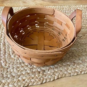 Può includere: Un cesto intrecciato con manici in pelle. Il cesto è fatto di legno marrone naturale e ha una forma rotonda. Il cesto è vuoto e pronto per essere riempito.