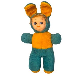 May include: A plush doll wearing a teal and orange bunny costume with long ears. The doll has a plastic face with blue eyes, light brown hair, and a small nose. The costume features a teal body and limbs with an orange chest panel and ear linings.
