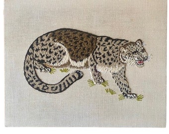 May include: A detailed embroidery of a snow leopard, rendered in shades of brown, black, and white, on a beige linen background. The leopard is depicted in a walking pose, with its head turned to the side, and its tail curled behind it.