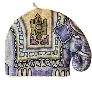 May include: A purple and beige embroidered tea cozy shaped like an elephant. The elephant has a detailed design with geometric patterns and a fringe around the bottom.