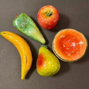 May include: A selection of colourful, handcrafted paper mache fruits. The assortment includes a yellow banana, a green pepper, a red apple, a green and yellow pear, and a watermelon slice. The fruits are arranged on a dark surface.