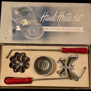 Op de afbeelding: Een vintage "Handi Hostess Kit" voor het maken van wafels en taartbodems. De set bevat drie metalen vormen: een bloem, een vlinder en een ronde vorm. De vormen bevinden zich in een witte doos met een blauw-witte illustratie van een damesschoen.
