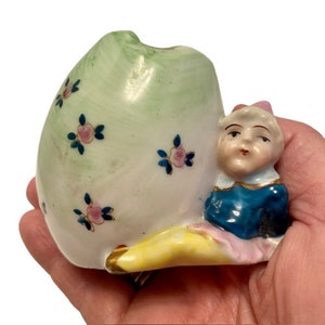 Könnte beinhalten: Eine Porzellanfigur eines Narren mit einer grün-weißen eiförmigen Vase. Der Narr trägt ein blaues und rosa Kostüm und einen gelben und weißen Hut. Die Vase ist mit rosa und blauen Blumen verziert.