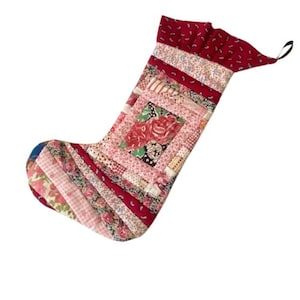 Puede incluir: Un calcetín de Navidad hecho a mano con un diseño de patchwork. El calcetín presenta una variedad de telas en tonos de rojo, rosa y blanco, con un cuadrado central que muestra un estampado floral. Se adjunta un lazo negro para colgar.