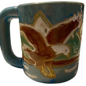 Könnte beinhalten: Eine türkisfarbene Keramiktasse mit großem Henkel. Die Tasse zeigt ein detailliertes Reliefdesign eines braunen und weißen Adlers im Flug vor einem grün-weißen Hintergrund. Die Tasse hat eine glänzende Oberfläche.