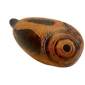 Puede incluir: Una calabaza tallada a mano con un diseño marrón y naranja. La calabaza tiene forma de pez y tiene un ojo grande tallado.