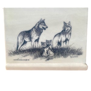 May include: A white marble plaque with a black and white drawing of a wolf family. The mother wolf is standing on the left side of the image, with two pups standing in front of her. The father wolf is standing on the right side of the image. The plaque is signed "Barrick" in the bottom right corner and "Montana Marble Inc. 1990" in the bottom left corner.