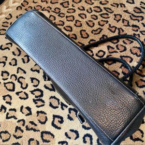 K&ouml;nnte beinhalten: Schwarze Ledertasche mit geflochtenem Trageriemen. Die Tasche hat eine strukturierte Oberfl&auml;che und eine wei&szlig;e Steppnaht an den R&auml;ndern.