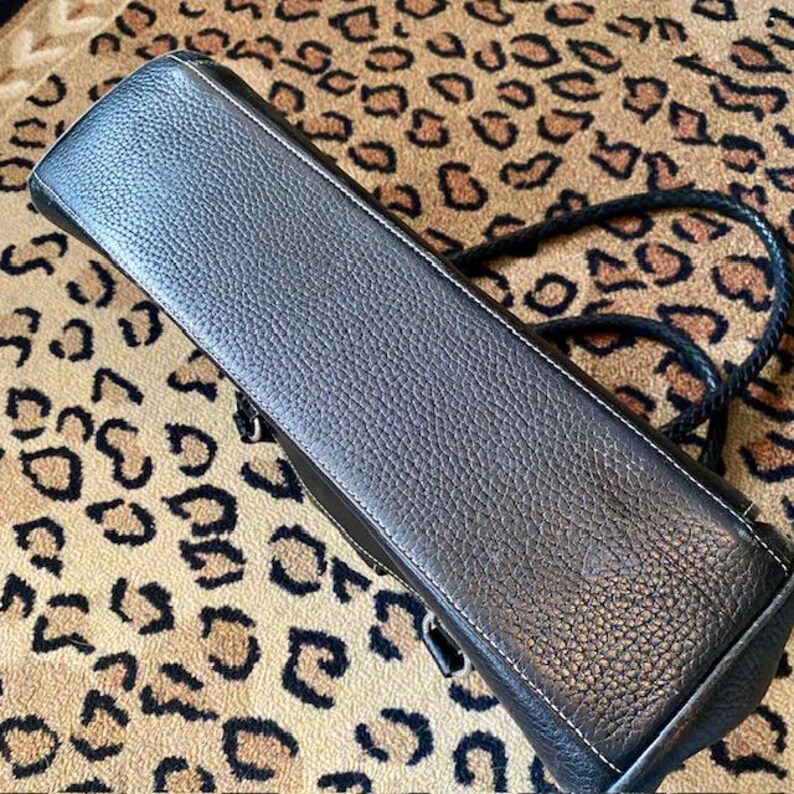 K&ouml;nnte beinhalten: Schwarze Ledertasche mit geflochtenem Trageriemen. Die Tasche hat eine strukturierte Oberfl&auml;che und eine wei&szlig;e Steppnaht an den R&auml;ndern.