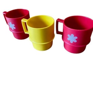 Puede incluir: Tres tazas de plástico de colores. Dos son rojas con un diseño de copo de nieve azul, y una es amarilla. Cada taza tiene un asa y una base apilada. Las tazas están vacías y listas para ser llenadas.