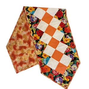 May include: A table runner with a checkered pattern in white and orange. The runner is bordered with a black fabric featuring a floral design with red, yellow, and green flowers and vintage record labels.