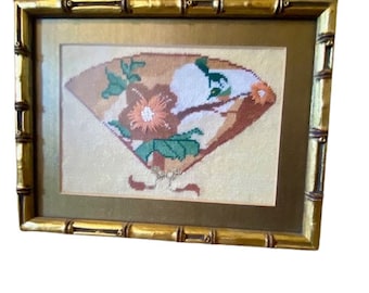 May include: A framed needlepoint embroidery of a fan with floral designs in shades of brown, green, and white. The frame is made of gold-painted bamboo.