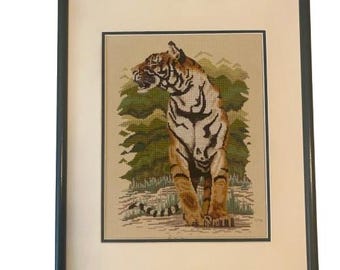 May include: Framed needlepoint art featuring a tiger. The tiger is depicted with orange and black stripes, set against a backdrop of green trees and a light beige background. The artwork is framed in a dark green wooden frame.