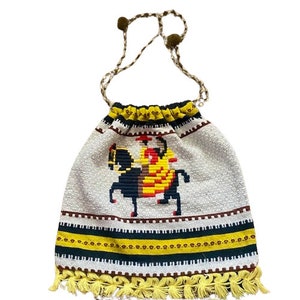 Puede incluir: Una bolsa de cordón de crochet blanco con un diseño bordado en amarillo, negro y rojo de una persona montando a caballo. La bolsa tiene un ribete de flecos y un asa de cordón marrón.