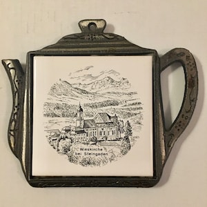 Puede incluir: Un salvamanteles decorativo con forma de tetera. Tiene un marco de metal y una inserción de baldosa cerámica blanca. La baldosa presenta una ilustración en blanco y negro de una iglesia y montañas, con el texto "Wieskirche bei Steingaden".