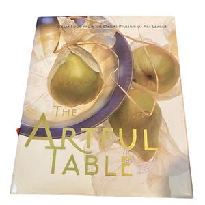 以下が含まれることがあります： 「The Artful Table」というタイトルの料理本。金色の文字が特徴です。表紙には、青い縁の皿にクッキングペーパーで包まれた梨が描かれています。「Great Food From The Dallas Museum of Art League」というテキストが上部にあります。