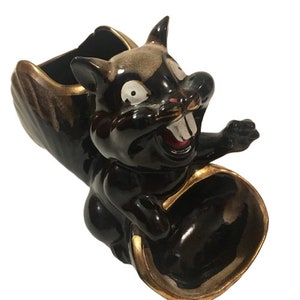 May include: A black and gold ceramic squirrel planter with a wide, open mouth and a gold rim. The squirrel is holding a small, round bowl in its paws.