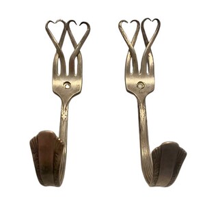 May include: Two silver fork hooks with heart-shaped tops. The forks are bent into a hook shape and the tines are twisted to form two hearts.