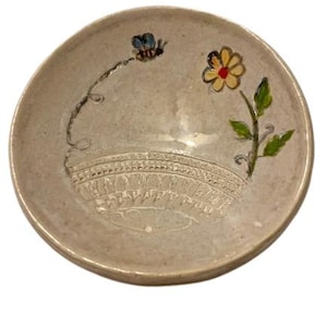 Puede incluir: Un plato de cerámica beige con un diseño pintado a mano. El plato presenta una abeja y una flor con pétalos amarillos y un centro rojo. El plato tiene un borde texturizado y un patrón decorativo en la parte inferior.
