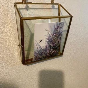May include: A gold-toned glass picture frame with a watercolor painting of a hummingbird flying near purple flowers. The frame is hanging on a white wall.