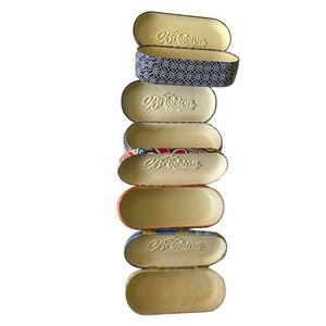 May include: A stack of seven eyeglass cases in various colors and patterns. The cases are oval-shaped with a hinged opening. The cases have a beige interior and a decorative exterior, including a case with a blue and white geometric pattern.