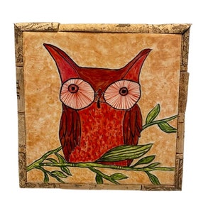 May include: A red owl with large, pink eyes sits on a branch with green leaves. The owl is painted on a corkboard with a light orange background.